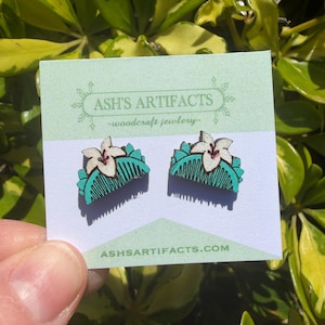 May include: A pair of wooden earrings featuring a white flower design atop a teal comb shape. The earrings are displayed on a light green card with the text "ASH'S ARTIFACTS - woodcraft jewlery" and the website address. The earrings are held in front of a green leafy background.