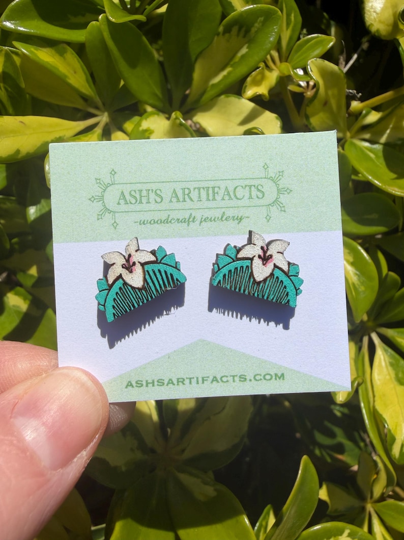 May include: A pair of wooden earrings featuring a white flower design atop a teal comb shape. The earrings are displayed on a light green card with the text "ASH'S ARTIFACTS - woodcraft jewlery" and the website address. The earrings are held in front of a green leafy background.