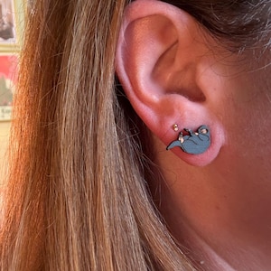 May include: A close-up of an ear with a blue otter-shaped earring. The wooden earring has a small red accent. The person's long, light brown hair is visible. The earring is a unique accessory.