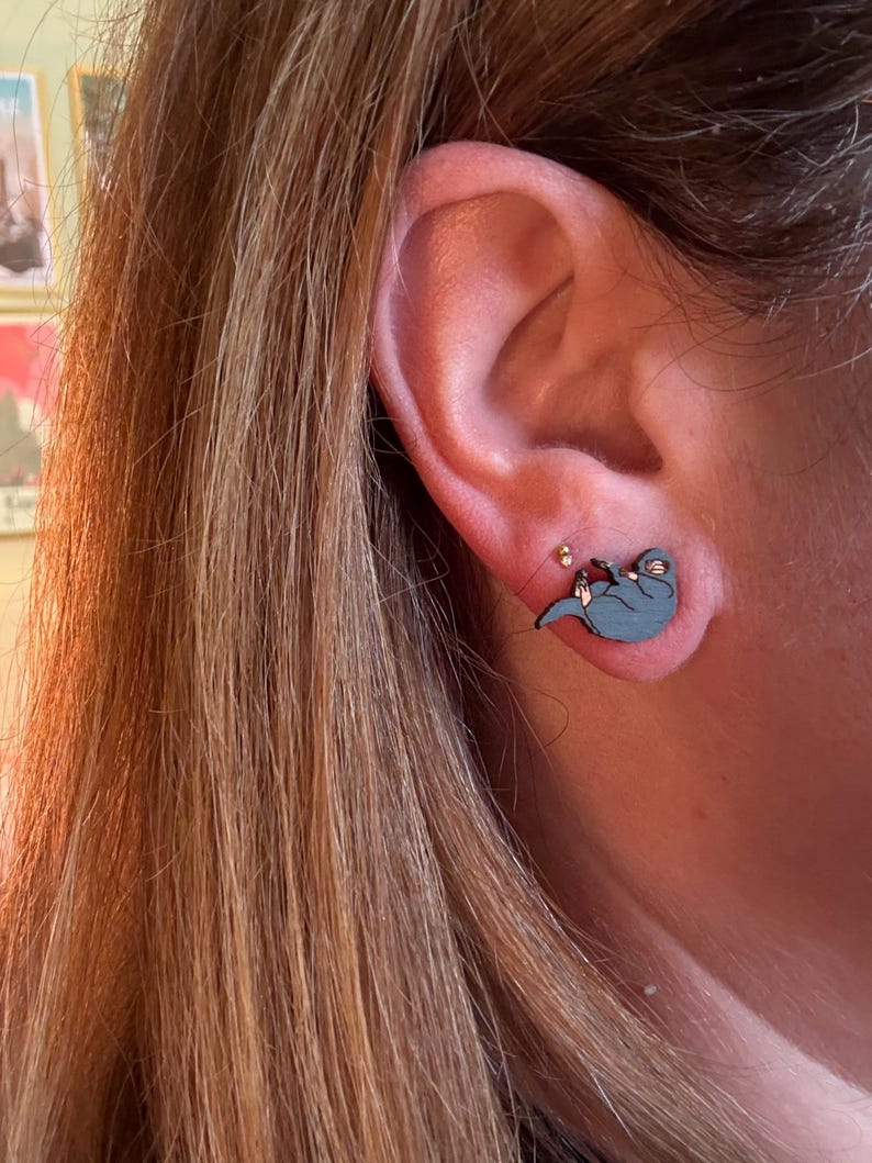 May include: A close-up of an ear with a blue otter-shaped earring. The wooden earring has a small red accent. The person's long, light brown hair is visible. The earring is a unique accessory.