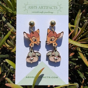 May include: Wooden dangle earrings featuring Bambi and Thumper characters. The earrings have gold-colored ball studs and are made of light brown wood. The earrings are displayed on a white card with the text "ASH'S ARTIFACTS - woodcraft jewelry" and the website address.