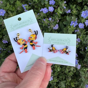 May include: Two pairs of wooden earrings featuring cartoon bees. The first pair features two bees with red and yellow bodies and black stripes. The second pair features two bees with yellow bodies and black stripes holding pink flowers. The cards are labelled "Ash's Artifacts - Woodcraft Jewellery" and "AshsArtifacts.com".