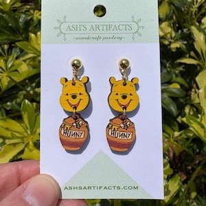 May include: Wooden earrings featuring Winnie the Pooh with a honey pot. The earrings are brown and yellow with gold accents. The honey pot has the word "Hunny" on it.