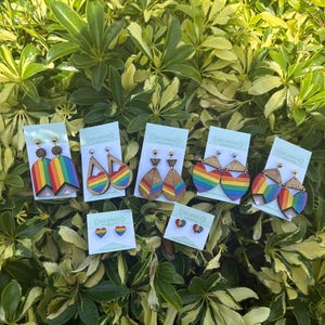 May include: A selection of wooden earrings featuring rainbow designs, including heart, teardrop, and geometric shapes. The earrings are presented on cards against a backdrop of green leaves.