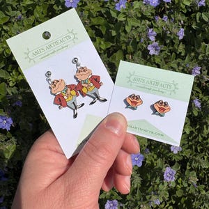 May include: Two pairs of wooden earrings featuring cartoon frogs wearing suits. The earrings are on white cards with the text "Ash's Artifacts - woodcraft jewelry" and the website "ashsartifacts.com".
