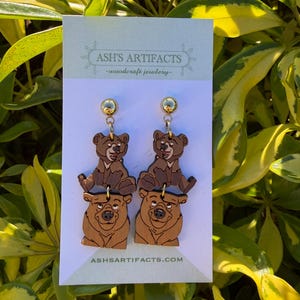 May include: Wooden bear earrings featuring a cartoon bear design. The earrings have a gold-colored stud and a two-tiered design with a bear cub on top of a larger bear. The earrings are displayed on a white card with the text "ASH'S ARTIFACTS -woodcraft jewlery-" and "ASHSARTIFACTS.COM".