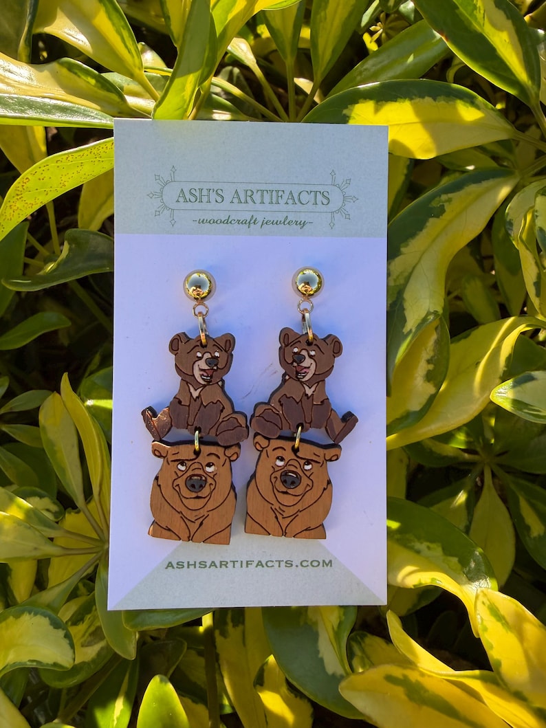 May include: Wooden bear earrings featuring a cartoon bear design. The earrings have a gold-colored stud and a two-tiered design with a bear cub on top of a larger bear. The earrings are displayed on a white card with the text "ASH'S ARTIFACTS -woodcraft jewlery-" and "ASHSARTIFACTS.COM".