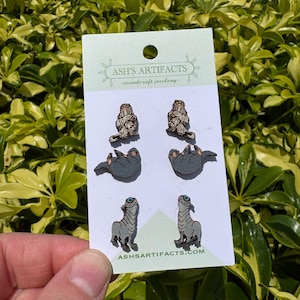May include: A card of woodcraft jewellery featuring six pairs of earrings. The earrings depict a sloth, a creature with a long neck, and a creature with a long neck. The card has the text "ASH'S ARTIFACTS - woodcraft jewellery" and "ASHARTIFACTS.COM".