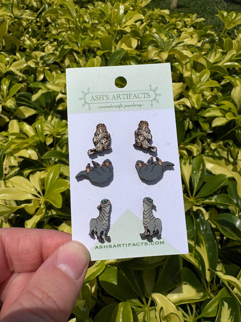 May include: A card of woodcraft jewellery featuring six pairs of earrings. The earrings depict a sloth, a creature with a long neck, and a creature with a long neck. The card has the text "ASH'S ARTIFACTS - woodcraft jewellery" and "ASHARTIFACTS.COM".