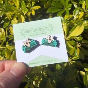 May include: Wooden earrings featuring a flower and comb design. The earrings are light brown, teal, and white, with a floral motif. The earrings are displayed on a light green card with the text "ASH'S ARTIFACTS - woodcraft jewelry" and "ashsartifacts.com".