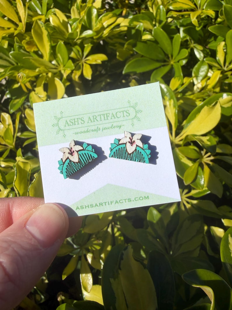 May include: Wooden earrings featuring a flower and comb design. The earrings are light brown, teal, and white, with a floral motif. The earrings are displayed on a light green card with the text "ASH'S ARTIFACTS - woodcraft jewelry" and "ashsartifacts.com".