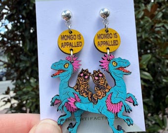 May include: A pair of dangle earrings featuring a yellow circular wooden charm with the text MONGO IS APPALLED, attached to a blue dinosaur figure with pink accents and two small brown furry creatures sitting on its back, on a display card.