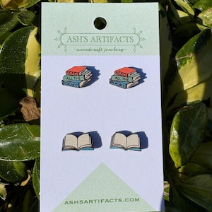 May include: A pair of wooden earrings featuring a stack of books with the text "Read All The Books" and a single open book. The earrings are on a white card with the text "Ash's Artifacts - woodcraft jewelry - ash'sartifacts.com".