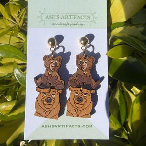 May include: Wooden bear earrings with gold-colored ball studs. The earrings feature a two-tiered design with a bear at the top and a larger bear at the bottom. The earrings are displayed on a white card with the text "ASH'S ARTIFACTS - woodcraft jewlery" and "ASHSARTIFACTS.COM".