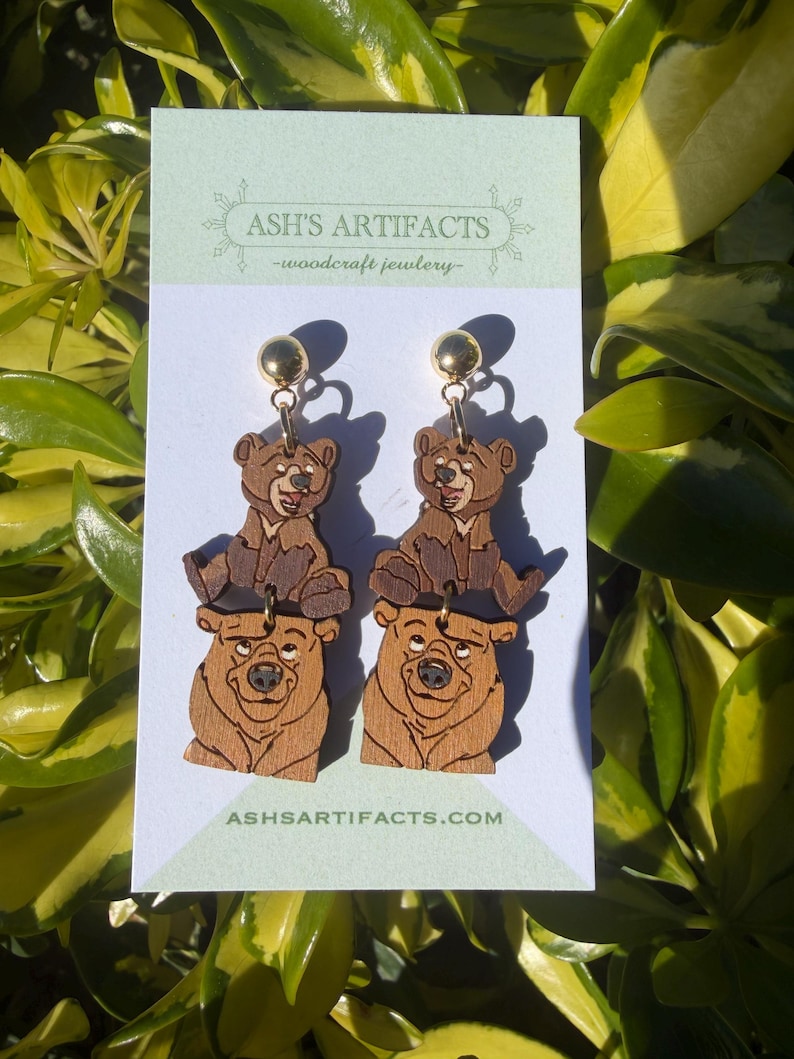 May include: Wooden bear earrings with gold-colored ball studs. The earrings feature a two-tiered design with a bear at the top and a larger bear at the bottom. The earrings are displayed on a white card with the text "ASH'S ARTIFACTS - woodcraft jewlery" and "ASHSARTIFACTS.COM".