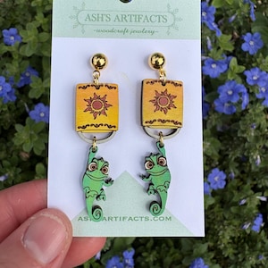 May include: A pair of wooden earrings featuring a green chameleon hanging from a yellow square with a sun design. The earrings are labeled "ASH'S ARTIFACTS - woodcraft jewelry - ASH'S ARTIFACTS.COM"