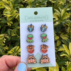 Puede incluir: Un conjunto de seis pendientes de madera con personajes de los Muppets. Los pendientes están en pares y presentan a Cookie Monster, Miss Piggy, Fozzie Bear y Kermit la Rana. Los pendientes están en una tarjeta blanca con el texto "ASH'S ARTIFACTS -woodcraft jewelry- ".