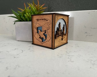 May include: A wooden box with a light brown stain and black trim. The box has a cutout of the Ravenclaw house crest from Harry Potter, with the words "Ravenclaw" above the crest. The box also has a cutout of Hogwarts castle on the side.