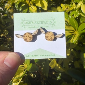 May include: Wooden earrings featuring a round design with wings, resembling a golden snitch. The earrings are displayed on a light green card with the text "ASH'S ARTIFACTS -woodcraft jewelry-" and the website address. The background is a lush green foliage.