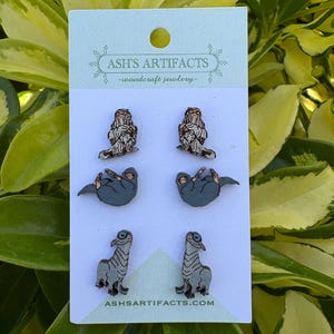 May include: A set of four pairs of wooden earrings with animal designs. The earrings include two pairs of sloths, two pairs of dinosaurs, and two pairs of furry creatures. The earrings are displayed on a white card with the text "ASH'S ARTIFACTS - woodcraft jewelry" and "ASHSARTIFACTS.COM".