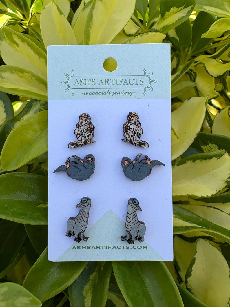 May include: A set of four pairs of wooden earrings with animal designs. The earrings include two pairs of sloths, two pairs of dinosaurs, and two pairs of furry creatures. The earrings are displayed on a white card with the text "ASH'S ARTIFACTS - woodcraft jewelry" and "ASHSARTIFACTS.COM".