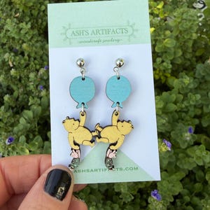 Puede incluir: Pendientes de madera con un diseño de globo azul claro y un personaje de oso amarillo colgando de él. Los pendientes se sujetan con un perno plateado. La tarjeta detrás dice "ASH'S ARTIFACTS - woodcraft jewelry - ash'sartifacts.com".