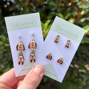 May include: Two sets of wooden earrings featuring the character Goofy on white display cards labeled ASH'S ARTIFACTS woodcraft jewelry. One set features dangling double-layered Goofy heads, while the other set consists of four individual stud earrings.