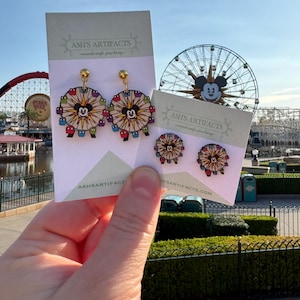 May include: A pair of earrings and a pair of stud earrings featuring a Ferris wheel design with a Mickey Mouse silhouette in the center. The earrings are on cards that read "ASH'S ARTIFACTS - woodcraft jewelry" and "ASH'S ARTIFACTS.COM".