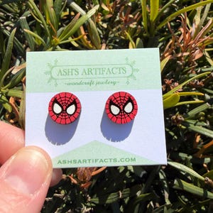 Puede incluir: Un par de pendientes redondos rojos y negros de Spider-Man con un diseño de telaraña blanca. Los pendientes se muestran en una tarjeta con el texto "ASH'S ARTIFACTS - woodcraft jewelry" y "ASHSARTIFACTS.COM".