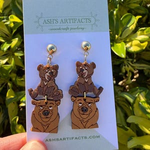 May include: Wooden bear earrings with gold-colored studs. The earrings feature a two-tiered design with a bear cub on top of a larger bear. The text "ASH'S ARTIFACTS -woodcraft jewelry-" is printed on the card. The website address is also printed on the card.