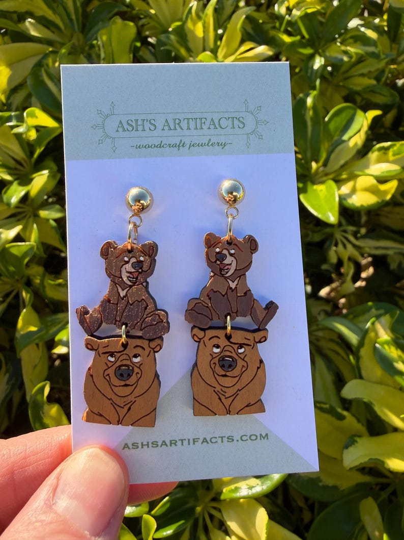 May include: Wooden bear earrings with gold-colored studs. The earrings feature a two-tiered design with a bear cub on top of a larger bear. The text "ASH'S ARTIFACTS -woodcraft jewelry-" is printed on the card. The website address is also printed on the card.