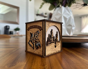 May include: A wooden box with a light brown finish. The box has a cutout of the Hogwarts crest on one side and a cutout of Hogwarts castle on the other side. The text "HOGWARTS" is visible on the crest.