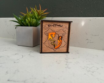 May include: A wooden box with a light brown finish and a black trim. The box has a cutout of the Gryffindor house crest, which is a lion with a red and gold shield. The text "Gryffindor" is written above the crest.