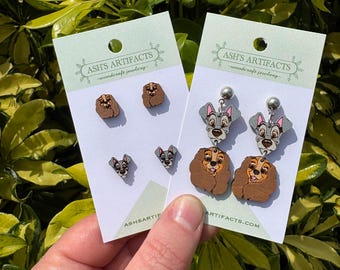 May include: Two pairs of wooden earrings featuring Lady and the Tramp characters. One pair features Lady and the other pair features Tramp. The earrings are on white cards with the text "Ash's Artifacts - woodcraft jewellery -" printed on them.