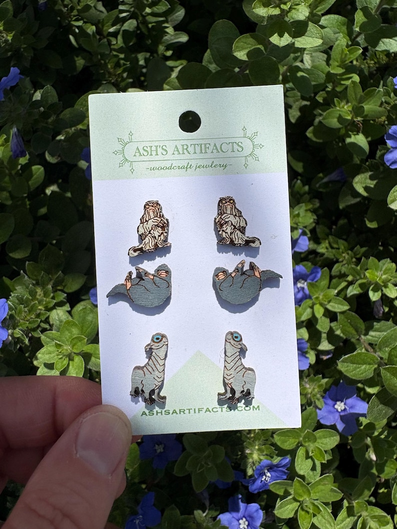 May include: A card of wooden earrings with three dinosaur designs. The card has the text "ASH'S ARTIFACTS - woodcraft jewelry" and a website address. The earrings are displayed against a backdrop of green foliage and blue flowers, typical of a garden.
