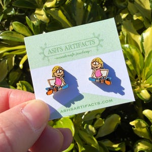 May include: A pair of wooden stud earrings featuring a cartoon girl sitting on a skateboard, wearing a pink top. The earrings are on a white card with the text "ASH'S ARTIFACTS - woodcraft jewelry" and "ASHSARTIFACTS.COM".