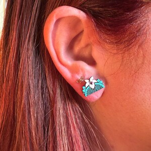 May include: A close-up of a small, wooden earring featuring a white flower and a turquoise comb design. The earring is attached to a gold-colored post. The earring is worn on a person's ear, with red hair visible in the background.