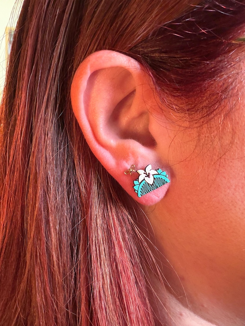 May include: A close-up of a small, wooden earring featuring a white flower and a turquoise comb design. The earring is attached to a gold-colored post. The earring is worn on a person's ear, with red hair visible in the background.