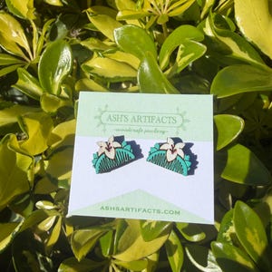 May include: A pair of wooden earrings featuring a comb design with a white flower and teal accents. The earrings are displayed on a white card with the text "ASH'S ARTIFACTS - woodcraft jewelry" and "ASHSARTIFACTS.COM". The background is a lush green and yellow foliage.
