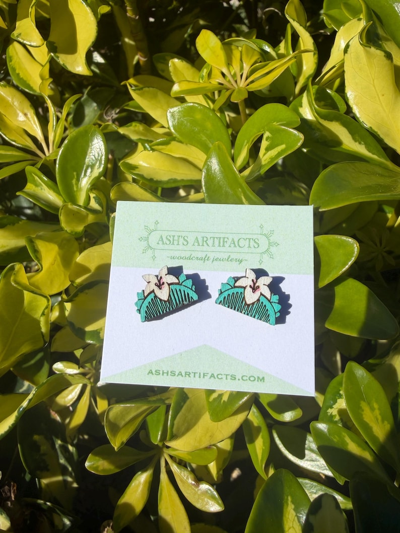 May include: A pair of wooden earrings featuring a comb design with a white flower and teal accents. The earrings are displayed on a white card with the text "ASH'S ARTIFACTS - woodcraft jewelry" and "ASHSARTIFACTS.COM". The background is a lush green and yellow foliage.