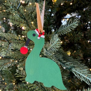May include: A green wooden dinosaur ornament with a red and white Santa hat. The dinosaur is holding a red ornament in its mouth. The ornament is hanging from a brown ribbon on a Christmas tree.
