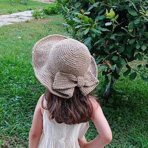 Op de afbeelding: Een bruine gehaakte zonnehoed met een grote strik. De hoed wordt gedragen door een persoon met lang bruin haar.