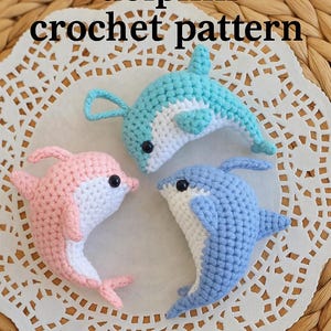 Puede incluir: Tres juguetes de delfines de ganchillo en tonos rosa, azul y celeste con vientres blancos. Cada delfín tiene ojos de seguridad negros. El texto "dolphin crochet pattern" está en la parte superior de la imagen.