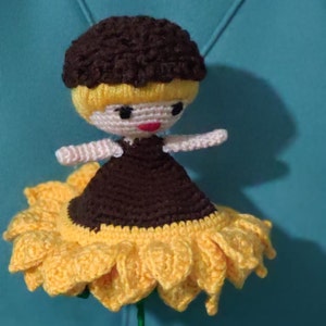 K&ouml;nnte beinhalten: Eine geh&auml;kelte Puppe mit braunen Haaren und einem schwarzen Kleid sitzt auf einer gelben geh&auml;kelten Sonnenblume.