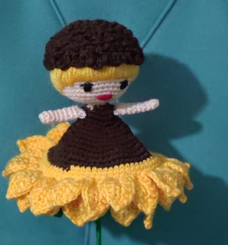 K&ouml;nnte beinhalten: Eine geh&auml;kelte Puppe mit braunen Haaren und einem schwarzen Kleid sitzt auf einer gelben geh&auml;kelten Sonnenblume.