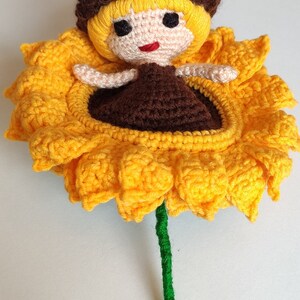 K&ouml;nnte beinhalten: Eine geh&auml;kelte Sonnenblume mit einer Puppe mit braunen Haaren, die darin sitzt. Die Sonnenblume ist gelb mit einem gr&uuml;nen Stiel.