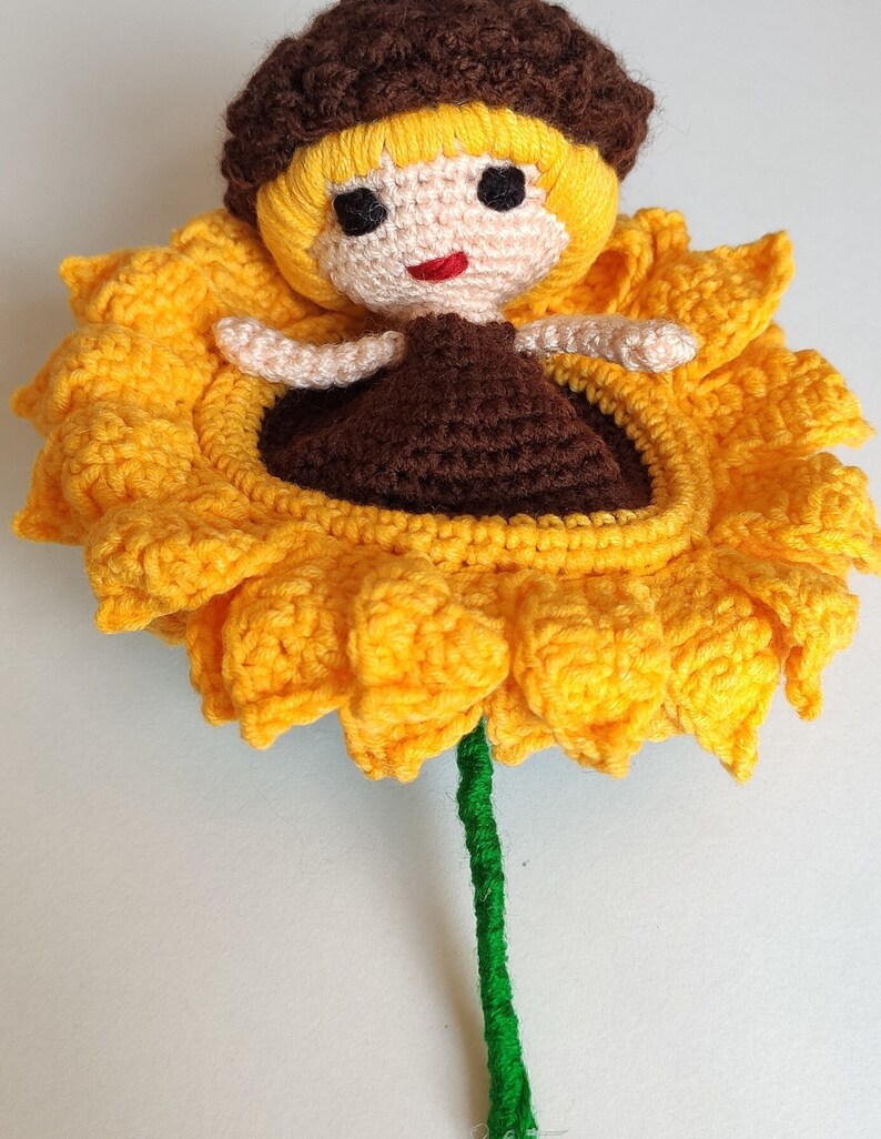 K&ouml;nnte beinhalten: Eine geh&auml;kelte Sonnenblume mit einer Puppe mit braunen Haaren, die darin sitzt. Die Sonnenblume ist gelb mit einem gr&uuml;nen Stiel.