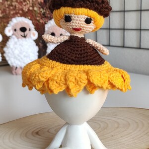 K&ouml;nnte beinhalten: Eine geh&auml;kelte Puppe mit braunen Haaren, einem gelben Kleid und einem Sonnenblumenhut. Die Puppe sitzt auf einem wei&szlig;en Sockel.