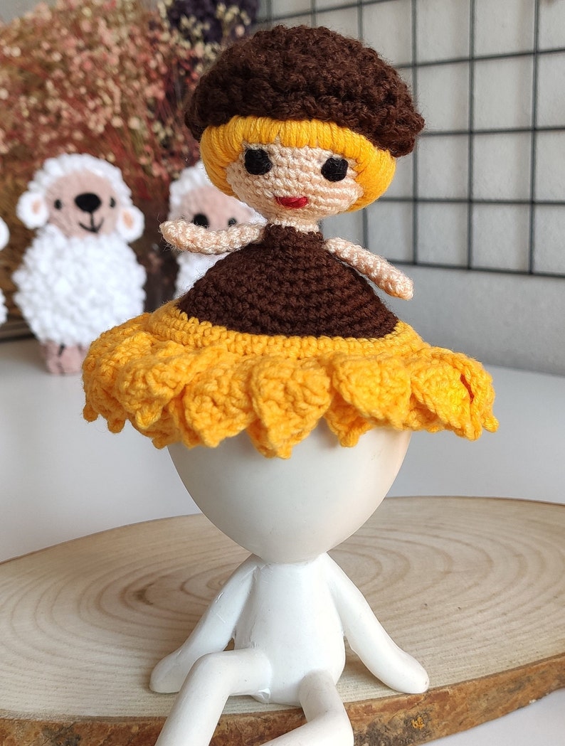 K&ouml;nnte beinhalten: Eine geh&auml;kelte Puppe mit braunen Haaren, einem gelben Kleid und einem Sonnenblumenhut. Die Puppe sitzt auf einem wei&szlig;en Sockel.