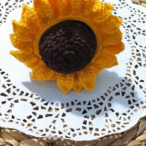 K&ouml;nnte beinhalten: Eine geh&auml;kelte Sonnenblume mit gelben Bl&uuml;tenbl&auml;ttern und einer braunen Mitte. Die Sonnenblume liegt auf einer wei&szlig;en Spitze.